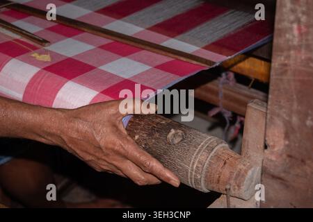 Nahaufnahme eines Handwerkers, der traditionellen Jamdani-Stoff in Rot-weiß-Karomuster auf einem hölzernen Handwebstuhl webt und handgefertigte Textilien zeigt. Stockfoto