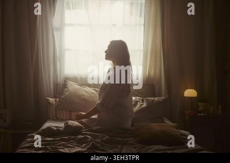 Junge Erwachsene Frau, die auf dem Bett sitzt und in der Nähe des Fensters meditiert, morgendliche Achtsamkeit und Stresslinderung übt. Nützlich für psychische Gesundheit, Selbstpflege, Wellness-Kampagnen Stockfoto