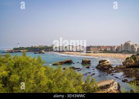 Atemberaubender Blick auf die Küste von Biarritz mit der überfüllten Grande Plage, zerklüfteten Felsen und dem berühmten weißen Leuchtturm in der Ferne. vordergrundgrün Stockfoto