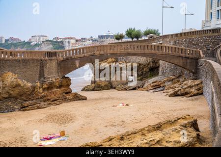 Eine malerische Steinbogenbrücke überspannt den Sandstrand in Biarritz, Frankreich. Die elegante Stadtarchitektur und der Leuchtturm bilden den Hintergrund. Eine Person Rela Stockfoto