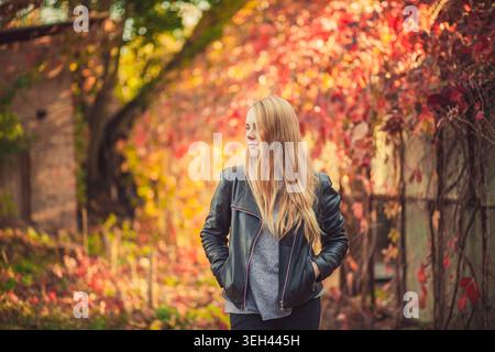 Eine junge Frau mit langen blonden Haaren in schwarzer Lederjacke steht nachdenklich im Herbstpark. Warmes Sonnenlicht beleuchtet die Szene mit Rot und Orange Stockfoto