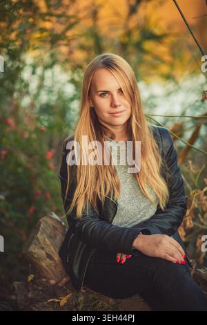 Junge Frau mit langen blonden Haaren sitzt im Herbst. Sie trägt eine schwarze Lederjacke und hat rote Nägel. Weiches natürliches Licht und warme Herbstfarben cre Stockfoto