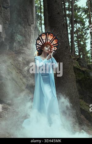 Mystische Frau in blauem Kleid im rauchigen Wald, filmisches Fantasy-Porträt mit surrealer Atmosphäre. Stockfoto