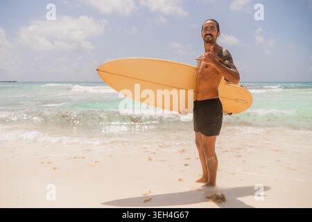 Energiegeladener, tätowierter Surfer, der an einem hellen Sandstrand steht und ein altes gelbes Surfbrett hält, während er in die Kamera lächelt. Stockfoto