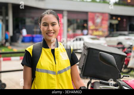 Ein horizontales mittleres Porträt fängt eine glückliche junge Isan Thai weibliche Zustellbotin ein, die während eines o mit einem fröhlichen Lächeln direkt in die Kamera blickt Stockfoto