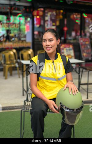 Ein vertikales Medium Shot fängt eine glückliche junge Isan Thai-Kurierin ein, die während einer Pause von ihrer Schicht auf einem Stuhl sitzt. Sie wir Stockfoto