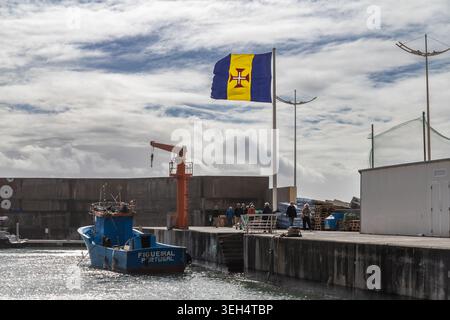 Portugal, Madeira, Calheta-2. Februar 2026: Die regionale Flagge Madeiras weht über einem kleinen Fischerhafen mit Booten, Dock-Infrastruktur und bewölktem Atlan Stockfoto