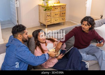Drei Freunde sitzen auf einem grauen Sofa im Wohnzimmer und teilen Popcorn aus einer klaren Glasschale Stockfoto