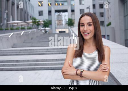 Selbstbewusste junge Frau, die draußen in der modernen Stadt mit überkreuzten Armen steht und vor dem Hintergrund der städtischen Architektur in die Kamera lächelt Stockfoto