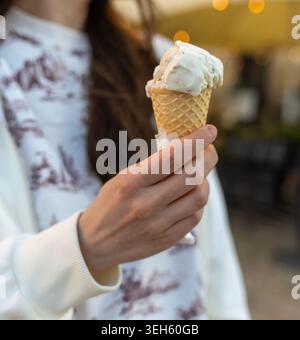 Eine Frau, die eine Eiskugel mit verschwommenen Hintergrundlichtern hält. Nahaufnahme des cremigen Desserts in Waffelkegel Stockfoto