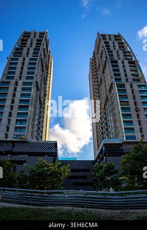 Flacher Blick auf zwei moderne Wohntürme, die sich in einen hellblauen Himmel mit Wolken erheben und eine beeindruckende urbane Architektur-Szene schaffen. Stockfoto