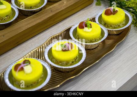 Eine Reihe eleganter, gelber Mini-Desserts, serviert in Folientassen auf einem dekorativen Tablett, garniert mit Obstgarnitur und Blumen, bereit für ein Buffet oder eine Feier Stockfoto