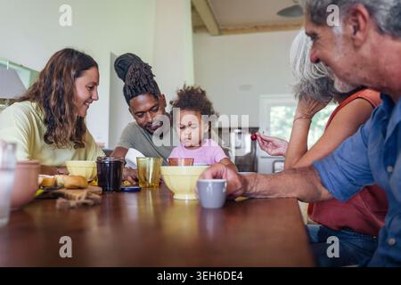 Multirassische Familie mit Großeltern, Eltern und Kleinkindern, die gemeinsam am Esstisch in gemütlicher Inneneinrichtung frühstücken. Stockfoto