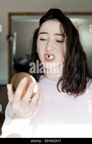 Eine reife Frau steht allein in ihrem Haus, hält einen Spiegel und drückt ihre Gefühle aus, dass ein Vorderzahn fehlt. Sie scheint über ihre Zahnarztpraxis nachzudenken Stockfoto