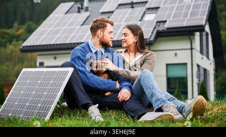 Paare sitzen auf Gras mit einem kleinen Hund in der Nähe eines modernen Hauses mit Sonnenkollektoren und schauen sich gegenseitig an. Photovoltaik-Dachanlage, erneuerbare Energien, umweltfreundlicher Lebensstil Stockfoto