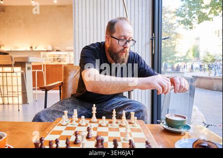 Bärtiger Mann, der langsam Getränke neben dem Spiel mischt. Gentleman mit Gesichtshaar rührt heißen Kaffee in ruhiger Umgebung. Ein bärtiger Mann konzentriert sich, während er seinen Kaffee in der Morgenruhe am Schachtisch vorsichtig rührt Stockfoto