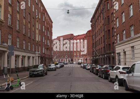 HELSINKI, FINNLAND - 11. AUGUST 2025: Breite Straße zwischen hohen Backsteingebäuden mit geparkten Autos und einem grünen Roller im Sommer. Stockfoto