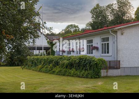 Weißes einstöckiges Haus mit roten Dachblumen und Fahnenmast hinter Hecke unter bewölktem Himmel. Stockfoto