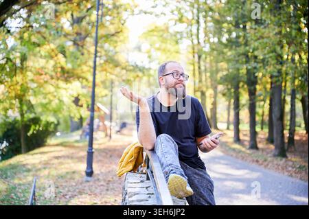 Ein Mann mit Bart zeigt Verwirrung beim Gehen. Ein bärtiger Mann scheint verwirrt zu sein, als er draußen mit dem Handy in der Hand schlendert. Lässig gekleideter Mann mit Bart zeigt Verwirrung auf Baumwänden im Freien Stockfoto