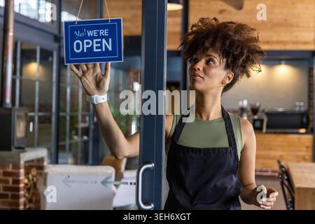 Afroamerikanerin, die ein blaues offenes Schild an der Glastür im Café mit schwarzer Schürze hängt Stockfoto