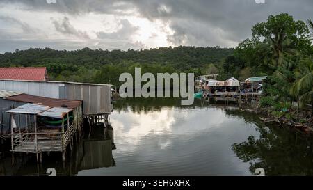 Ein ruhiges vietnamesisches Fischerdorf mit verwitterten Pfahlhäusern, die einen ruhigen Wasserweg säumen, flankiert von Mangroven und dichten Dschungelhügeln unter einem dramatischen Ov Stockfoto