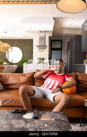 Mann, der auf braunem Sofa im Wohnzimmer sitzt, rotes Jersey trägt, Basketball hält und Glas schlürft Stockfoto