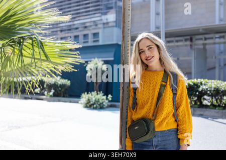 Frau 20, die sich an die Stange lehnt, lächelnd, mit Senfpullover, grüner Tasche, Palmwedel, Kopierbereich Stockfoto