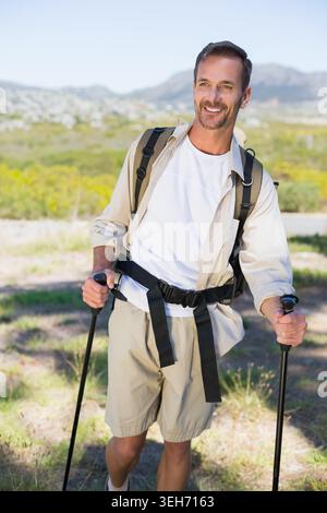 Mann mittleren Alters, der auf einem sonnendurchfluteten Naturpfad mit schwarzen Trekkingstöcken und Rucksack unterwegs ist Stockfoto