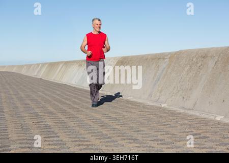 Senior Mann joggt entlang der gepflasterten Promenade neben einer schrägen Betonmauer, trägt rote Spitze Stockfoto