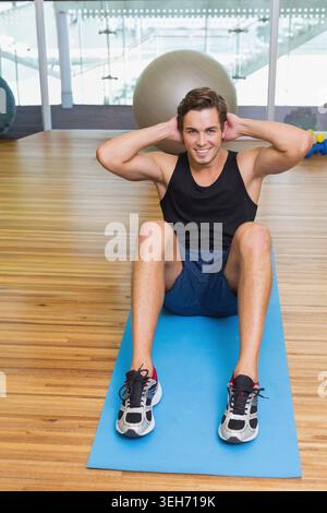 Mann sitzt auf blauer Matte im Fitnessstudio, lächelnd, trägt ein schwarzes ärmelloses Top, einen Stabilitätsball dahinter Stockfoto