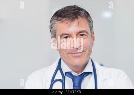 Lächelnd und in die Kamera schauend, reifer männlicher Arzt mit Labormantel, blauer Krawatte, Stethoskop in der Klinik Stockfoto