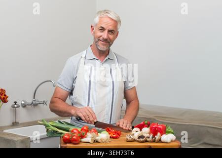 Reifer Mann, der Gemüse auf einem Schneidebrett an der Theke hackt und eine Schürze trägt, die das Messer des Küchenchefs hält Stockfoto