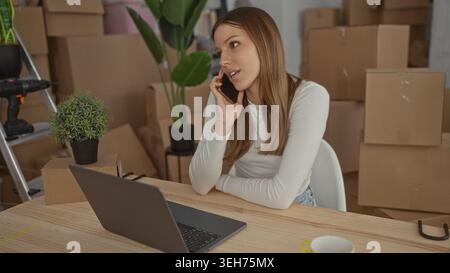 Junge Frau, die das Telefon am Ohr hält und auf einem Laptop in gestapelten Umzugskästen im Gebäude schreibt; Stress. Stockfoto