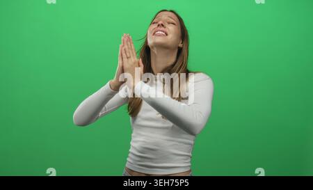 Junge hispanische Frau mit zusammengedrückten Händen betend im grünen Atelier, Augen geschlossen und ein leichtes Lächeln; Hoffnung. Stockfoto