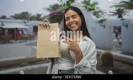 Die junge Frau lächelt und zeigt mit dem Finger auf eine braune Papiertüte, während sie sie in einer belebten Fußgängerzone in der Brust hält; fröhliche Beförderung. Stockfoto