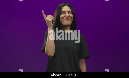 Frau zeigt den Zeigefinger nach oben in Richtung Tempel im Studio mit violettem Hintergrund und dezentem Lächeln; Inspiration für Selbstvertrauen. Stockfoto