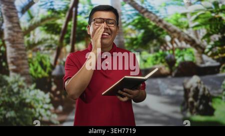 Mann liest aus dem Buch mit Hand-zu-Mund-Geste im Waldpark mit rotem Polo und Brille; Freude beim Lesen. Stockfoto