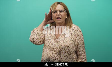 Frau, die Hand an Ohr schröpft und hinter einer Brille in einem Studio-blaugrünen Hintergrund knirscht; Hörschwierigkeiten sind besorgniserregend. Stockfoto