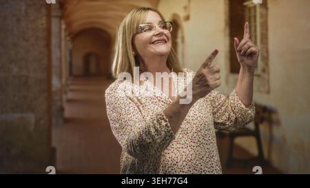 Die Frau zeigt die Zeigefinger mit lächelndem Gesicht und einer Brille in einem historischen Gebäudeflur nach oben, während sie mit den Händen nach oben blickt; Nostalgie Freude. Stockfoto