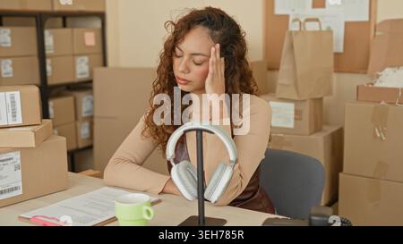 Junge hispanische Frau mit Hand zum Tempel, die Paketpapiere neben gestapelten Versandkartons im Gebäude liest; Stressbestandsmanagement. Stockfoto
