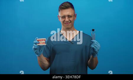 Männlicher Zahnarzt junger blonder in blauen Peelings und Handschuhen, die Zahnmodell und Spiegelwerkzeug mit Handschuhen halten, grimmig während Brille im Atelier; d Stockfoto