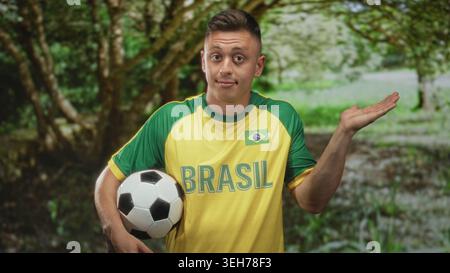 Ein Mann, der Fußball mit der Hand nach oben im Wald hält und brasilianisches Trikot mit Flagge auf der Brust trägt, sieht unsicher aus; Ungewissheit bei der Fanunterstützung. Stockfoto