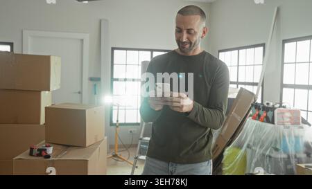 Mann überprüft Smartphone, Faustpumpen neben gestapelten Kartonschachteln und Leiter in einem Gebäude; Erleichterung Neuanfang. Stockfoto
