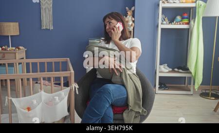 Frau, die das Smartphone am Ohr hält, während sie eine eingewickelte Babydecke neben einer hölzernen Krippe im Kinderzimmer hält; müdes Mitgefühl. Stockfoto
