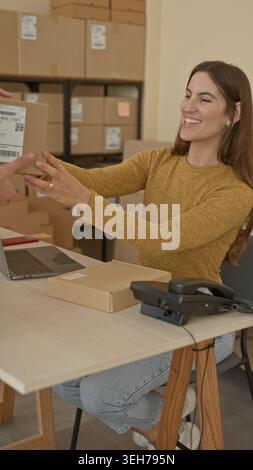 Frau, die ein Paket von einem Mann in einem kleinen Geschäftsgebäude erhält, Hände, die das Paket auf einem Schreibtisch mit Laptop und Versandkartons halten; Zusammenarbeit. Stockfoto