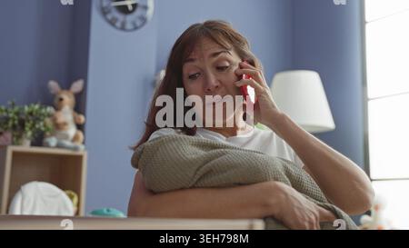 Die Frau hält das Smartphone an das Ohr und umarmt die Decke im Babyzimmer im Gebäude; Sorgen, Müdigkeit, Stress, überwältigt. Stockfoto