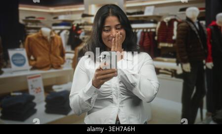 Frau lächelt und schickt auf dem Smartphone, wobei die Hände in einem Bekleidungsgeschäft zwischen Jacken und Schaufensterpuppen sichtbar sind; Einkaufsmöglichkeiten für Glücksgefühle. Stockfoto