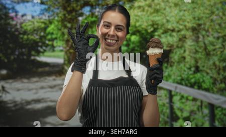 Frau auf Schürze, die Eisbecher hält und im Wald eine gute Geste macht, lächelt in die Kamera; fröhlicher Service. Stockfoto