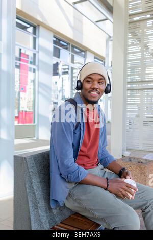 Erwachsener afroamerikanischer Mann, der auf einer Bank im Atrium sitzt, in Beanie und Kopfhörern, die eine Kaffeetasse halten Stockfoto