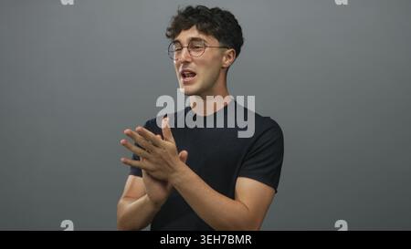 Junger Mann, der lächelnd die Hände klatscht, runde Brille und schwarzes T-Stück in einem grauen Studio trägt, kurze lockige Haare sichtbar; Freude feiern. Stockfoto